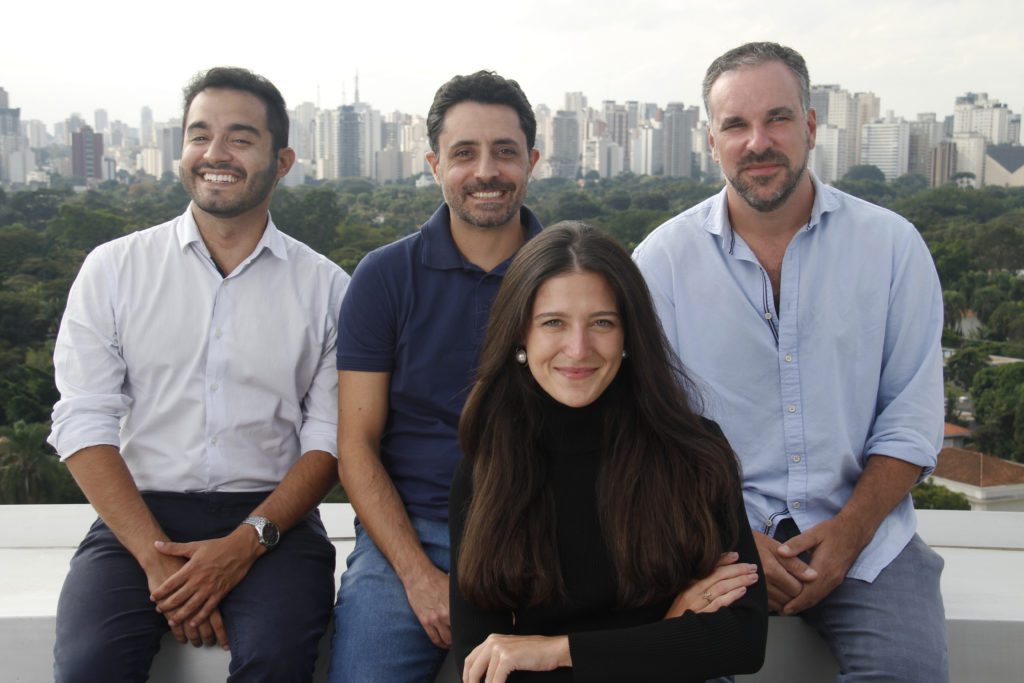 Quatro pessoas (uma mulher, três homens) sorrindo, sentadas em um terraço com vista para a cidade.
