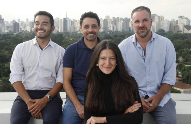 Quatro pessoas (uma mulher, três homens) sorrindo, sentadas em um terraço com vista para a cidade.