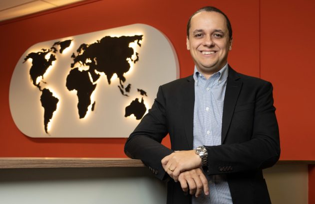 Homem sorridente de terno, apoiado em balcão, com mapa-múndi luminoso na parede.