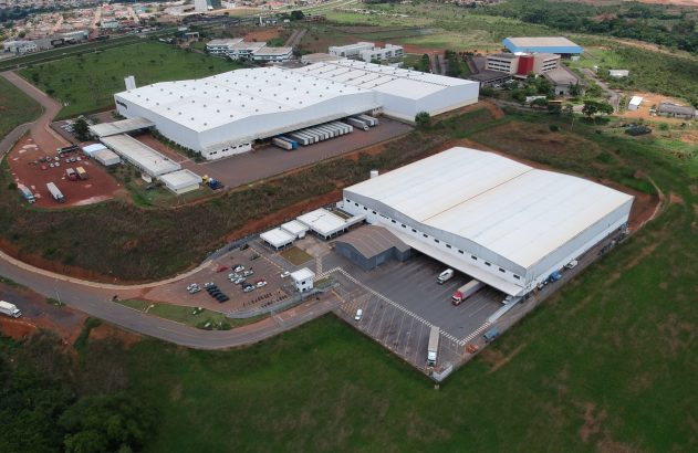 Vista aérea de complexo industrial com grandes galpões brancos e caminhões em movimentação.