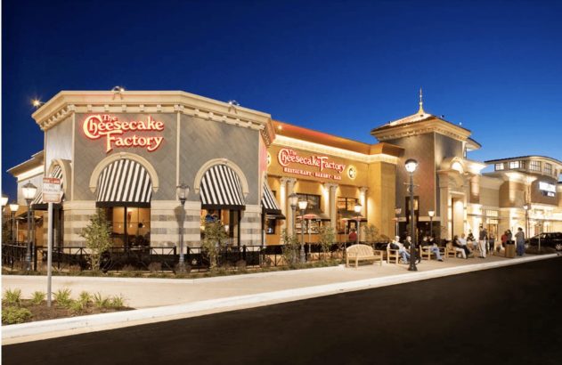 Restaurante The Cheesecake Factory à noite, com fachada iluminada, toldos listrados e pessoas sentadas em bancos externos.