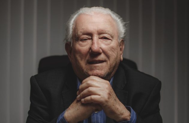 Homem sênior de cabelos brancos e terno escuro, com as mãos unidas sob o queixo, olhando diretamente.