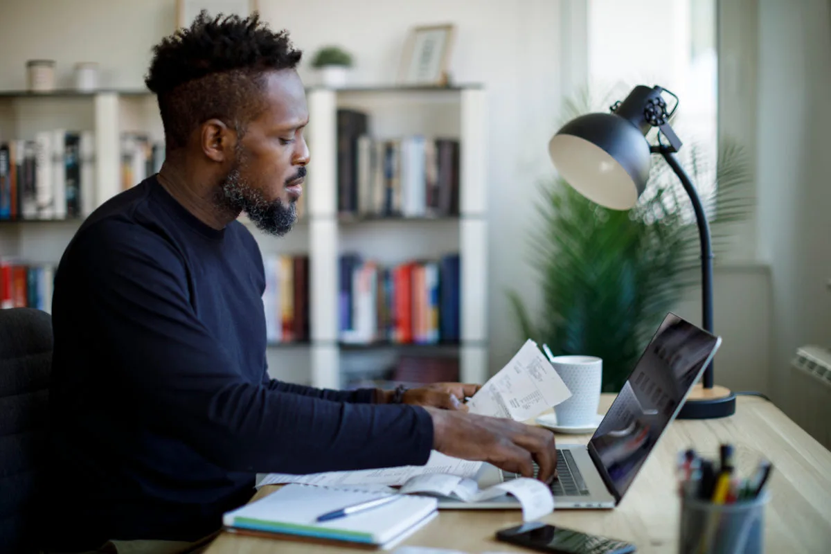 Homem negro teclando em um notebook com uma mão, enquanto a outra segura um documento que pode ser uma nota fiscal