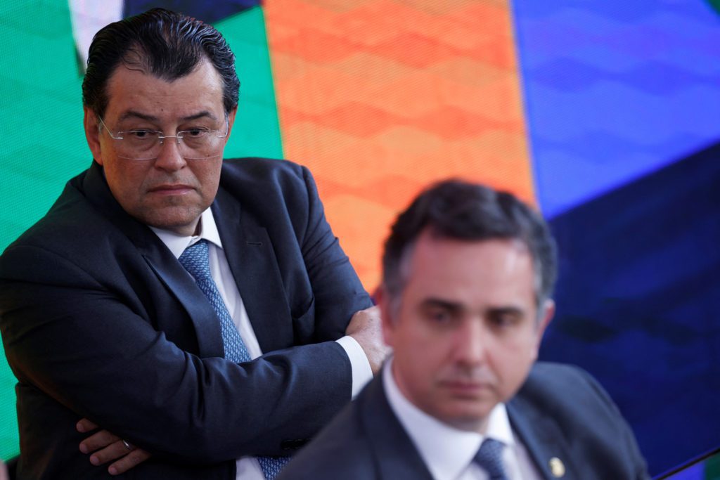 Homem de terno e óculos, braços cruzados, olhar sério. À direita, outro homem de terno desfocado.
