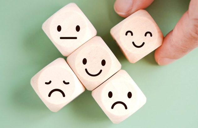 Mão posiciona cubo sorridente entre cubos de madeira com emoticons de emoções variadas.