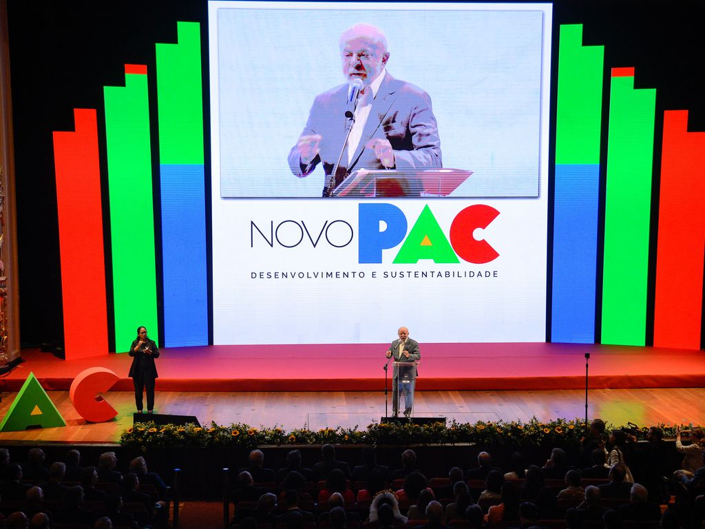 Palco de evento com tela LED exibindo "NOVO PAC" e um homem discursando. Um orador está no púlpito.