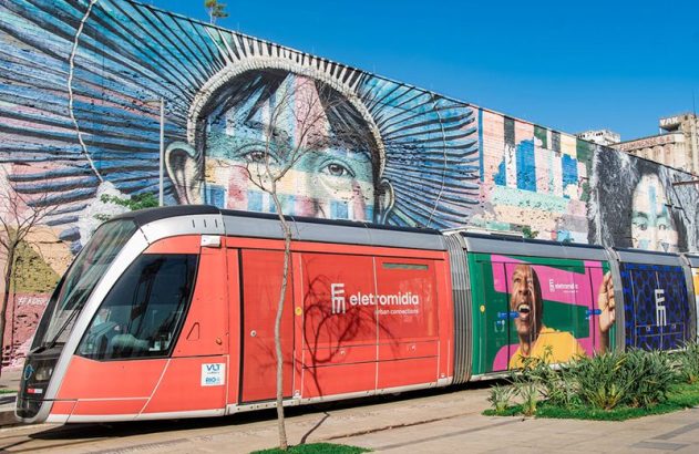 VLT vermelho e colorido parado em frente a um grande mural de rua vibrante sob o sol.
