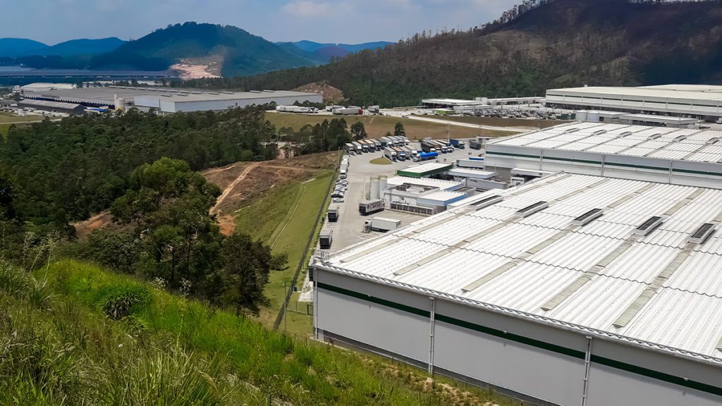 Grande complexo industrial com caminhões, rodeado por florestas, morros e um lago distante.