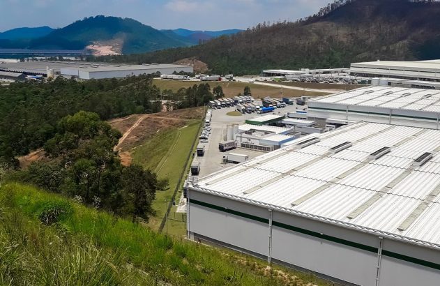 Grande complexo industrial com caminhões, rodeado por florestas, morros e um lago distante.