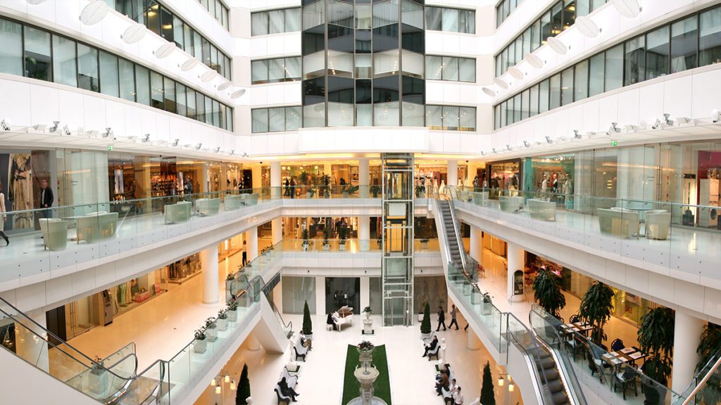 Vista aérea de shopping com vários níveis, pessoas, lojas, escadas rolantes e área central com piano e fonte.