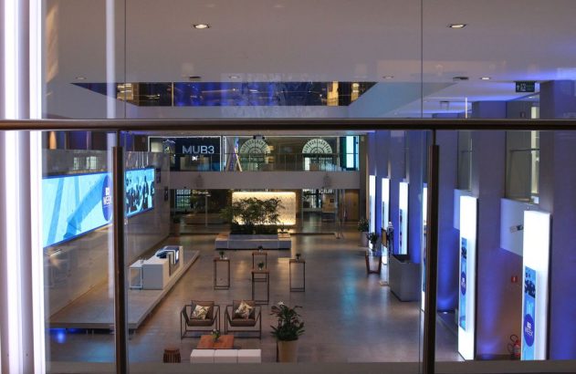 Saguão moderno com telas digitais e iluminação azul. Mobiliário e plantas decoram o ambiente visto de cima.