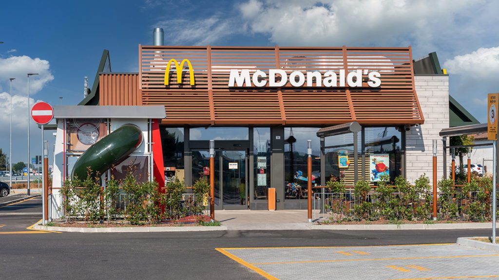 McDonald's moderno com fachada marrom, janelas amplas, drive-thru e um tubo verde na frente.