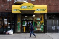 Fachada de loja Subway com marquise amarela e sinais "Open 24 Horas". Pessoa com mochila caminha na calçada.