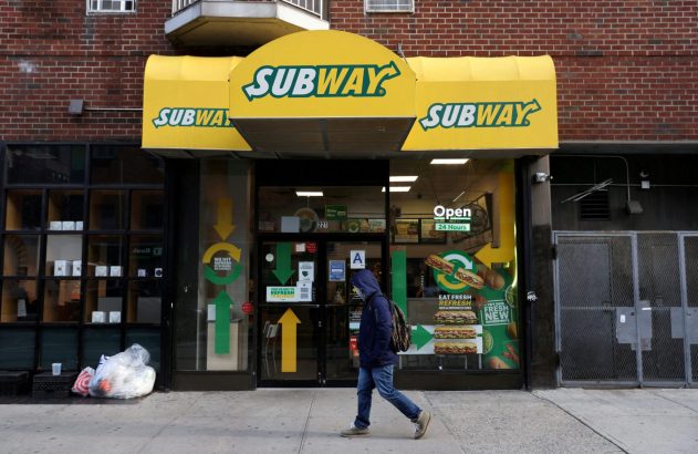Fachada de loja Subway com marquise amarela e sinais "Open 24 Horas". Pessoa com mochila caminha na calçada.