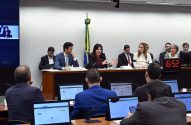 Reunião: Mulher fala em mesa principal, ladeada por outros. Plateia com laptops assiste em sala formal com bandeira.