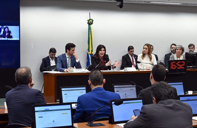 Reunião: Mulher fala em mesa principal, ladeada por outros. Plateia com laptops assiste em sala formal com bandeira.
