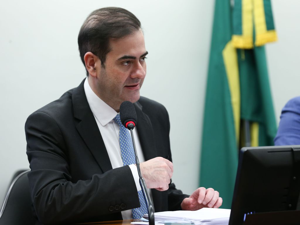 Homem de terno falando ao microfone em mesa. Bandeira do Brasil ao fundo. Ambiente oficial.