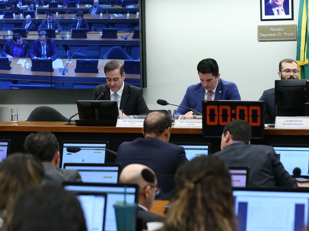 Sessão de plenário com deputados e secretários sentados, tela exibindo participantes e timer digital zerado.