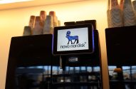 Máquina de café preta com tela exibindo o logo da Novo Nordisk, e copos empilhados no topo.