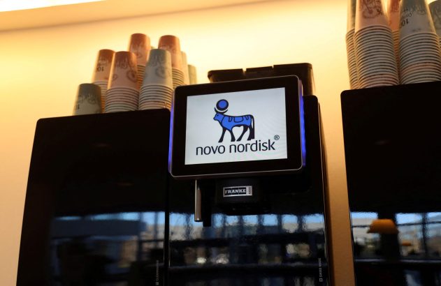 Máquina de café preta com tela exibindo o logo da Novo Nordisk, e copos empilhados no topo.