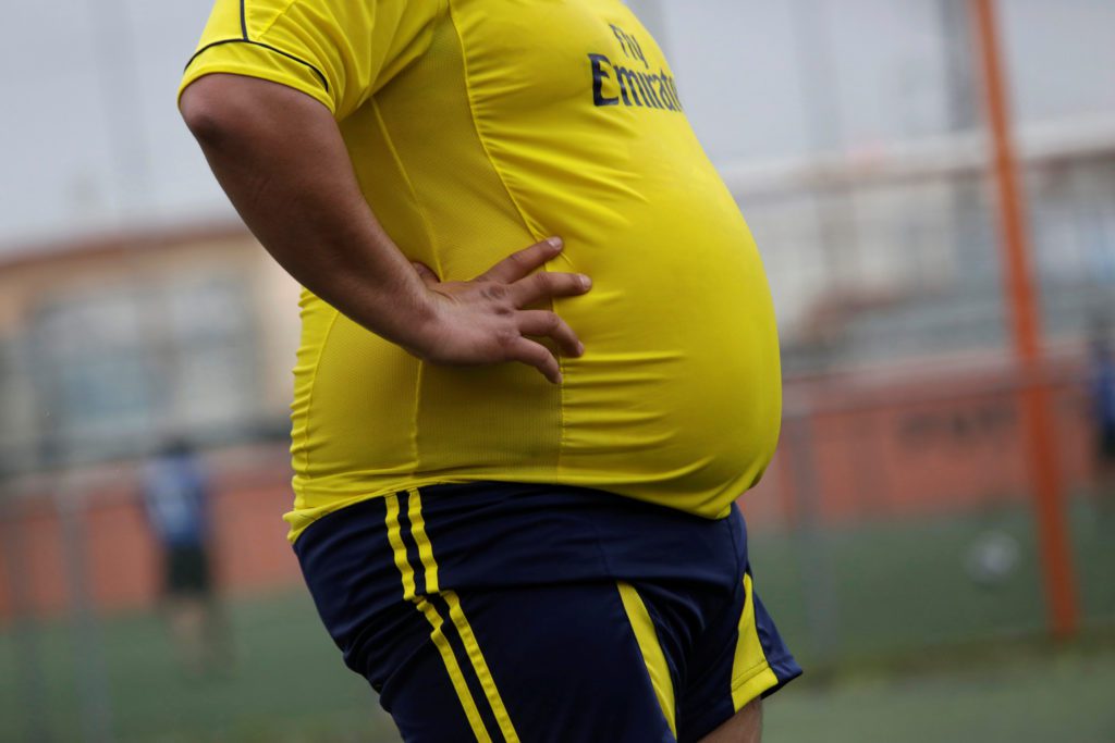 Pessoa com excesso de peso, camisa amarela justa, mãos na cintura, em campo de futebol.