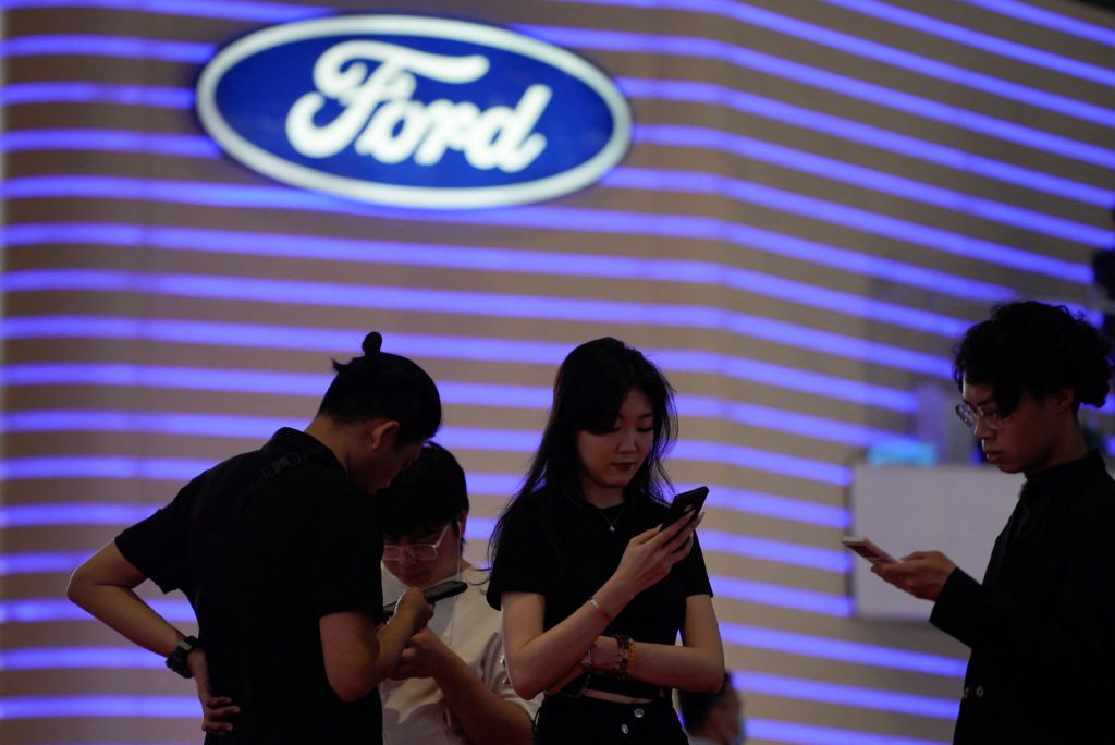 Quatro pessoas em frente ao logo da Ford, parede com luzes azuis, todas usando celulares.