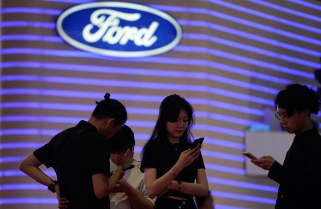 Quatro pessoas em frente ao logo da Ford, parede com luzes azuis, todas usando celulares.