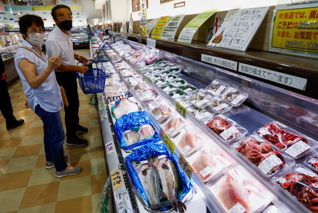 Casal de idosos de máscara em supermercado, observando vitrine refrigerada com frutos do mar embalados: peixes, polvos, mariscos.
