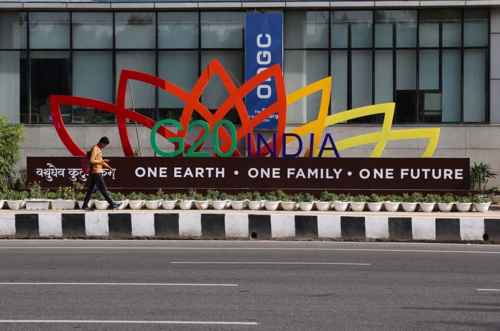 Letreiro do G20 Índia com "Uma Terra • Uma Família • Um Futuro". Um homem caminha ao lado, com prédio e rua ao fundo.