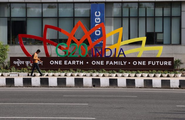 Letreiro do G20 Índia com "Uma Terra • Uma Família • Um Futuro". Um homem caminha ao lado, com prédio e rua ao fundo.