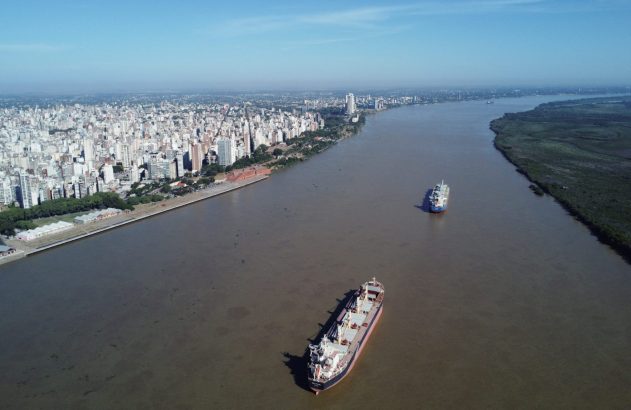 Vista aérea: Múltiplos navios cargueiros navegando em rio largo, cidade na margem esquerda e floresta na direita.