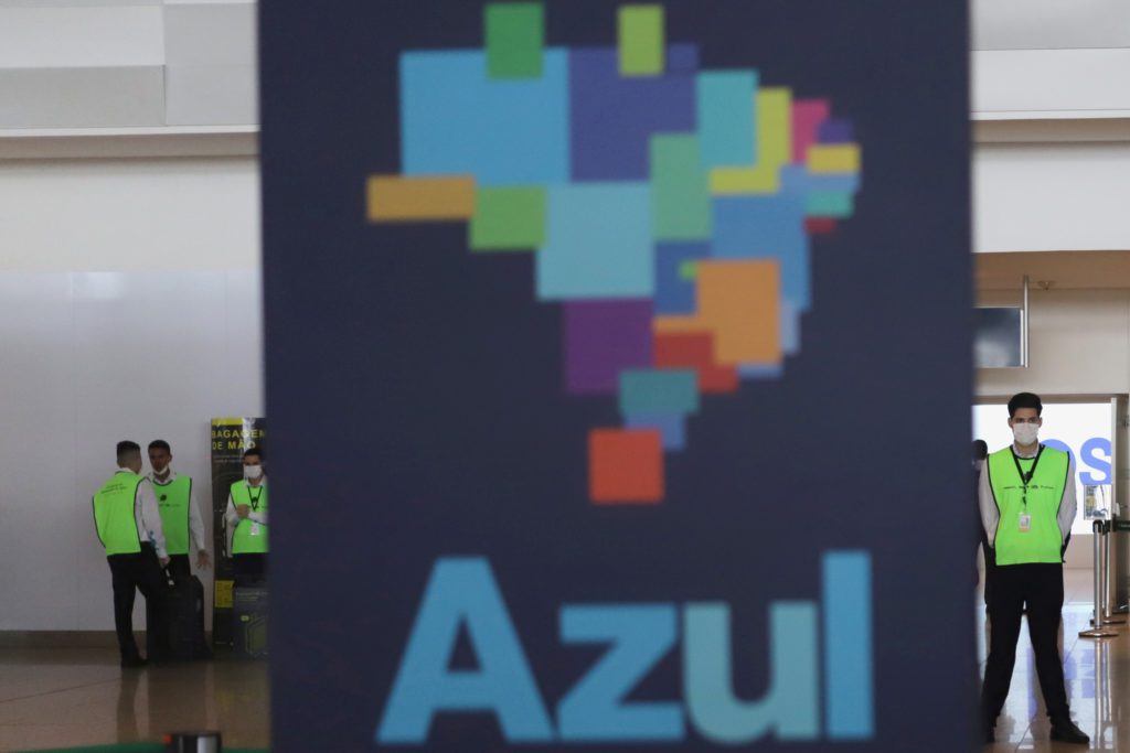 Homens de coletes verdes em aeroporto, atrás de um cartaz grande da Azul Linhas Aéreas com mapa do Brasil em blocos.