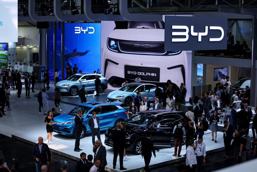 Estande da BYD em feira, com múltiplos carros em exposição. Pessoas observam veículos e telas exibem a marca.