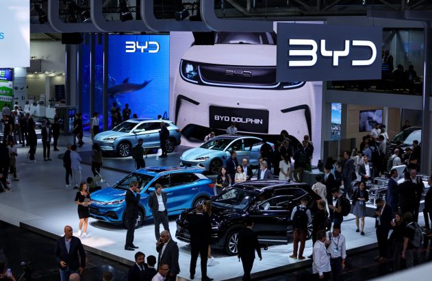 Estande da BYD em feira, com múltiplos carros em exposição. Pessoas observam veículos e telas exibem a marca.