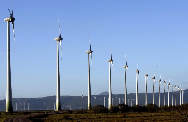 Aerogeradores em fileira num parque eólico, com montanhas e céu azul ao fundo.