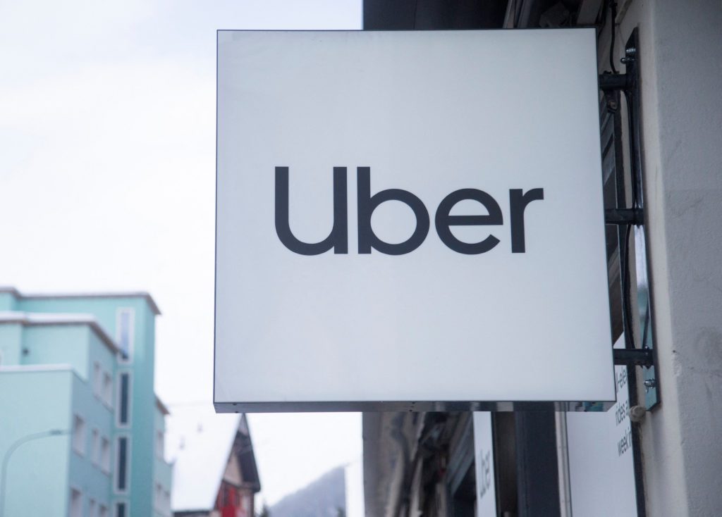 Letreiro branco da Uber pendurado em prédio, com outros edifícios ao fundo e céu claro.