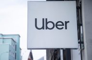 Letreiro branco da Uber pendurado em prédio, com outros edifícios ao fundo e céu claro.