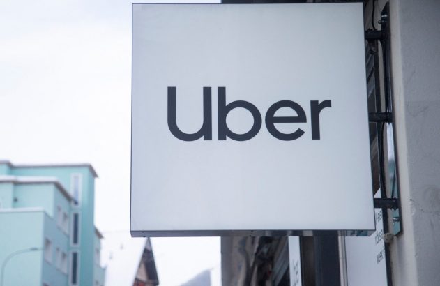 Letreiro branco da Uber pendurado em prédio, com outros edifícios ao fundo e céu claro.