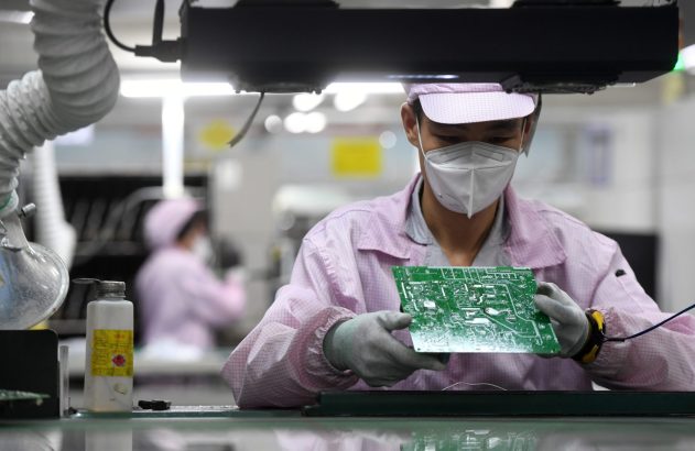 Operário com máscara e luvas inspeciona placa de circuito eletrônico em linha de montagem, outros operários ao fundo.