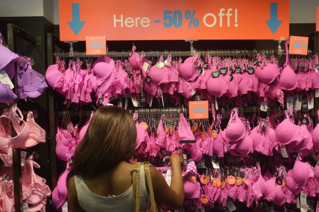 Pessoa de costas em loja de lingerie, olhando sutiãs rosa em araras com 50% de desconto.
