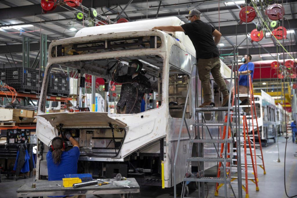 Funcionários montam a estrutura de um ônibus em uma fábrica. Um solda componentes, outro em escada.