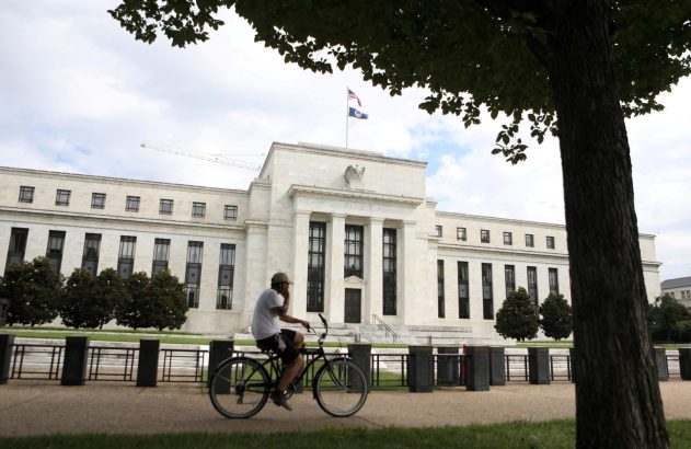 Ciclista em frente ao Federal Reserve, edifício branco com colunas e bandeiras dos EUA.