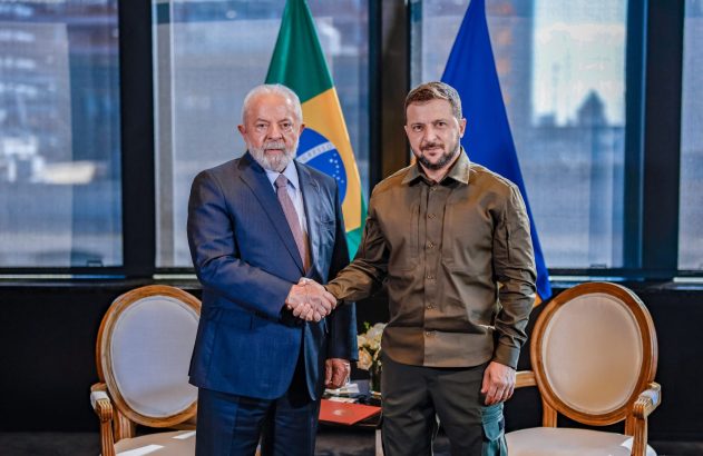Presidentes Lula (Brasil) e Zelenskyy (Ucrânia) apertam as mãos. Bandeiras ao fundo.
