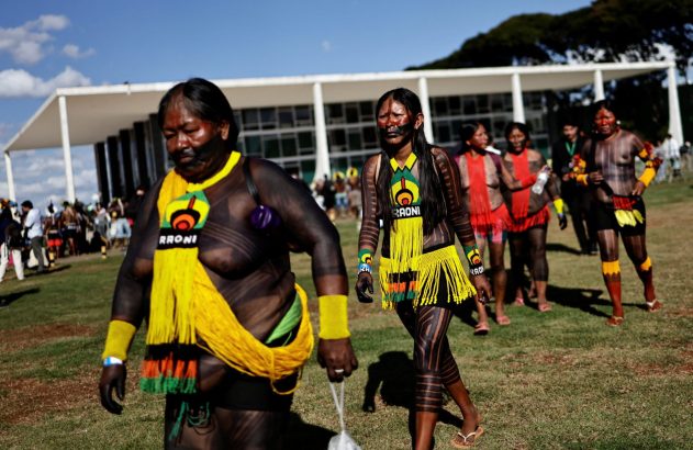Grupo de indígenas com vestimentas tradicionais e pinturas corporais caminha em frente a um edifício governamental.