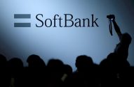 Logo SoftBank com símbolo de igual em painel iluminado. Público silhuetado e pessoa fotografando.