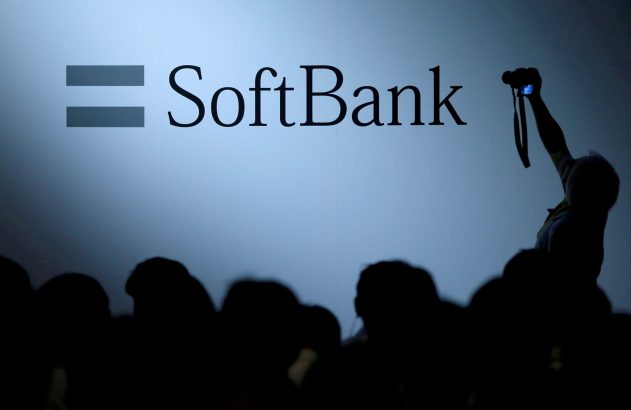 Logo SoftBank com símbolo de igual em painel iluminado. Público silhuetado e pessoa fotografando.