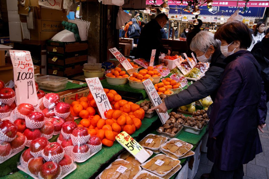 Duas mulheres com máscaras observam frutas e produtos frescos em uma banca de mercado. Maçãs e tangerinas em destaque. Uma aponta.