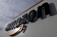 Logotipo da Amazon em preto e laranja, com a seta dourada, fixado em uma parede externa clara.