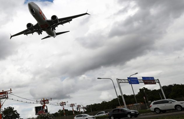 Avião em aproximação sobre rodovia com veículos e sinalização, balizamento de aeroporto e céu nublado.