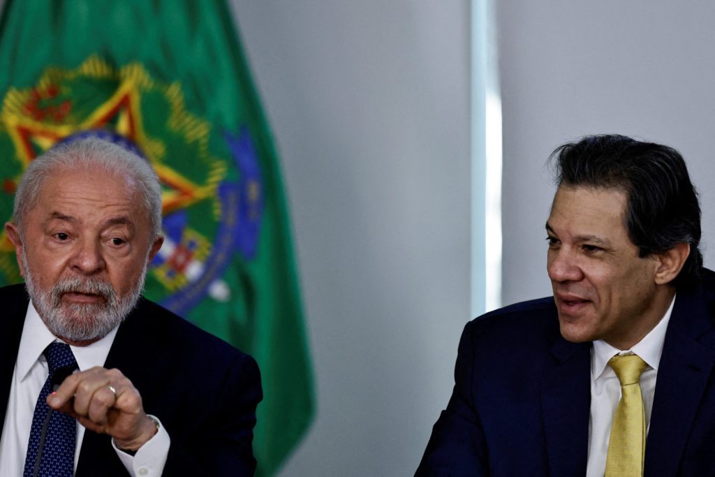 Lula gesticula para Haddad, que ouve atento, em reunião. Bandeira brasileira ao fundo.